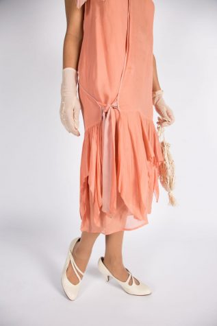 Details on the coral chiffon dress with multi-tiered relaxed ruffle skirt, bow detail and a asymmetric hemline. Accessorized with white heels and mid-length gloves.
