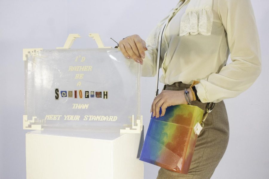 Clear bag that looks like a briefcase has white text on it saying I'd rather be a sociopath than meet your standard. The word sociopath is with magazine cut outs of letters that very in color but match the size of the rest of the text. A person is leaning next to the bag with a rainbow crossbody on. 