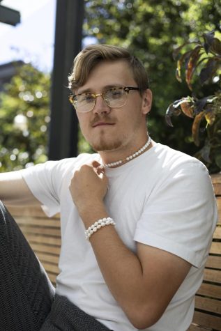 Man sitting on bench outside, a bush is behind him, he has one had on his knee that is up and the other is grabbing his pearl necklace