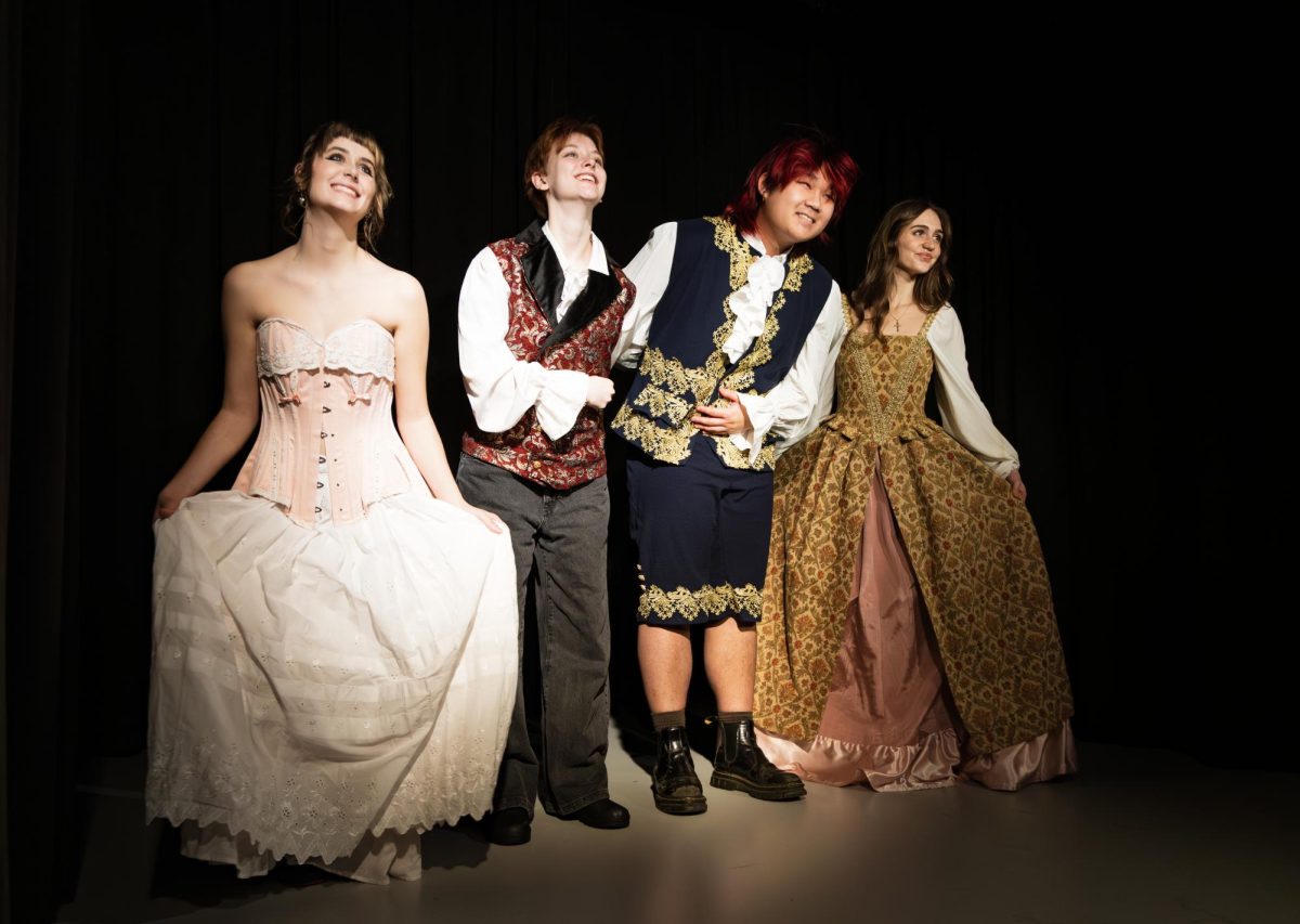 Four people stand together on a stage under spotlight lighting, wearing theatrical historical-style costumes including corsets, embroidered vests, and long dresses against a red curtain backdrop.